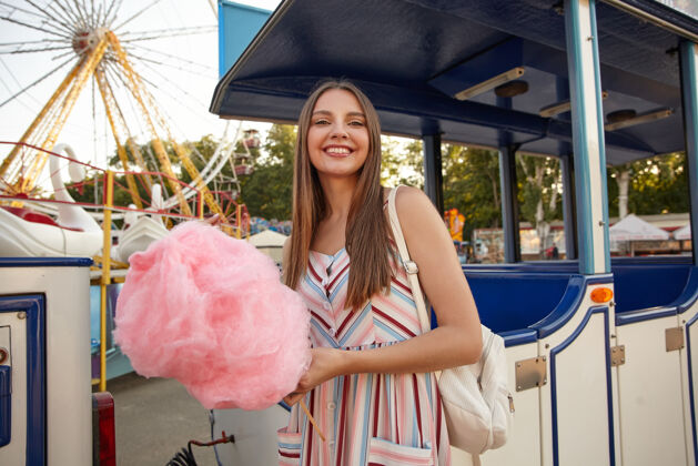 衣服阳光明媚的日子里 年轻迷人的棕发女性在游乐园里摆着姿势 穿着浪漫的连衣裙和背包 手里拿着粉色的棉花糖头发休闲年轻