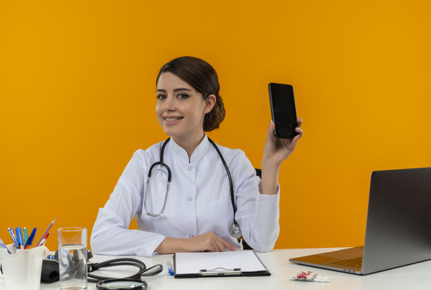 工作微笑的年轻女医生穿着医用长袍 手持听诊器 坐在办公桌旁 拿着医疗工具 拿着手机 背景是黄色的医生女听诊器