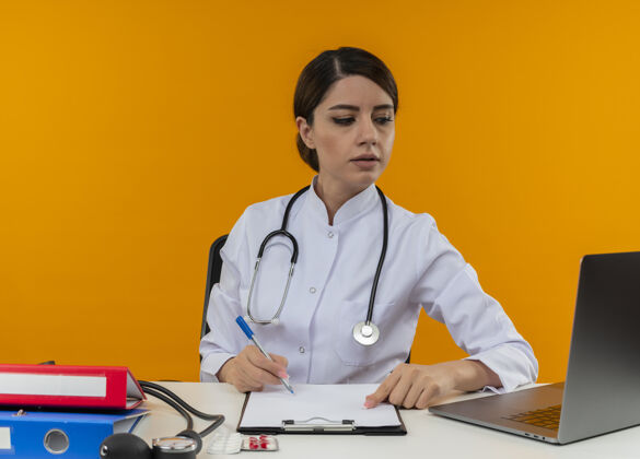 电脑想着年轻的女医生穿着医用长袍带着听诊器坐在办公桌前拿着医疗工具坐在电脑上看隔离黄色背景上的笔记本电脑工作医疗思考