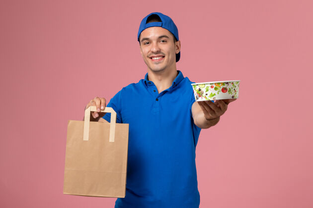 递送正面图：身穿蓝色制服和斗篷的年轻男性快递员 手上拿着快递包裹和碗 放在粉红色的墙上制服工作快乐