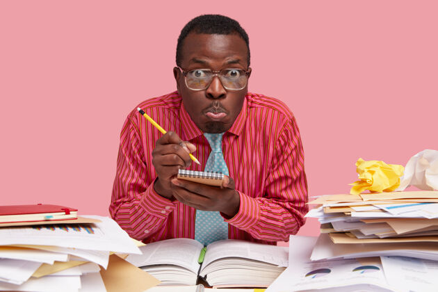 桌面情绪激动的黑人横拍在笔记本上写下信息 独自坐在桌面上 做鬼脸 穿着粉色衬衫和领带 在报告上工作眼镜努力工作文书