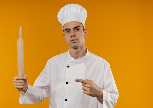 制服穿着厨师制服的年轻男子用手指着他手上的擀面杖 站在隔离的黄色墙壁上手滚点