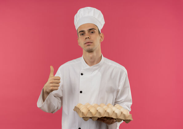 持有自信的年轻男厨师穿着厨师制服 拿着一批鸡蛋 大拇指竖立在孤立的粉红色墙上鸡蛋拇指厨师