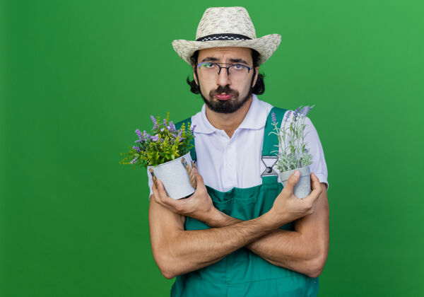 悲伤年轻的留着胡子的园丁 穿着连体衣 戴着帽子 手里拿着盆栽植物 表情悲伤胡须园丁年轻