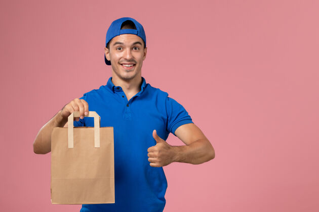 送货正面图：身穿蓝色制服和披风的年轻男性快递员 手上拿着一个纸包裹 放在粉红色的墙上人男人快乐