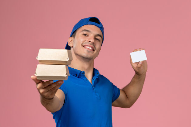 人正面图：身穿蓝色制服披肩的年轻男性快递员拿着小包裹 浅粉色墙上有卡片卡片男性粉色