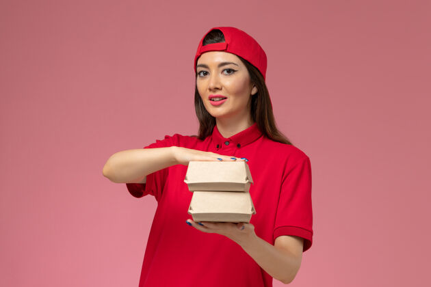 快乐正面图：身穿红色制服和斗篷的年轻女快递员 手上拿着小包裹 放在粉红色的墙上年轻女快递服务手