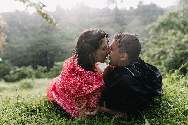 棕榈树穿着粉色雨衣的性感女人在旅行中亲吻她的男朋友一对浪漫的旅行者躺在草地上的户外照片青春男性感性
