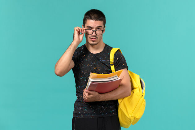 学院身穿深色t恤黄色背包的男学生正面图 浅蓝色的墙上放着文件和书籍教训微笑男学生