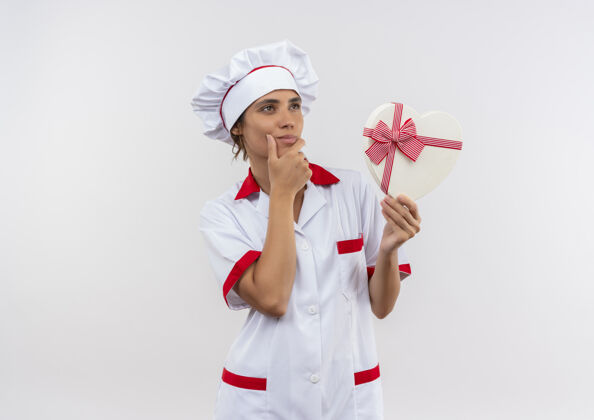 穿看着一旁想着年轻的女厨师穿着厨师制服拿着心形盒子把手放在下巴上孤立的白色墙壁上留着临摹的空间侧女性下巴