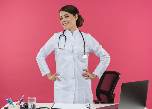 电脑微笑的年轻女医生穿着医用长袍 手持听诊器站在办公桌后面 手持医疗工具 双手放在臀部 留有复印空间医疗桌子放
