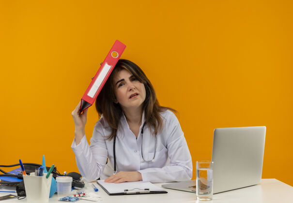 坐一位穿着医用长袍 手持听诊器的中年女医生坐在办公桌旁 拿着医疗工具在笔记本电脑上工作 把文件夹放在头上 放在隔离的橙色墙上 留有复印空间医生文件夹笔记本电脑